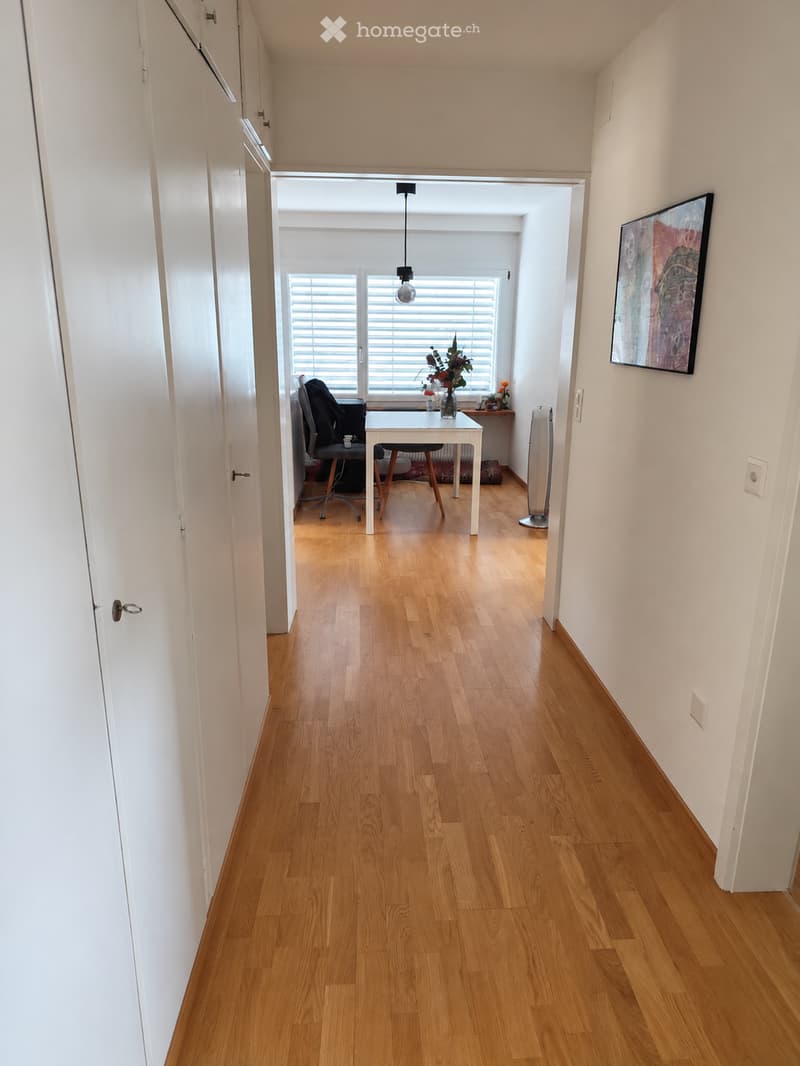 A long, narrow hallway with light wooden flooring and white walls. The hallway leads to a room with a desk, chair, and a vase with flowers on a small table. There is a painting on the wall to the right.