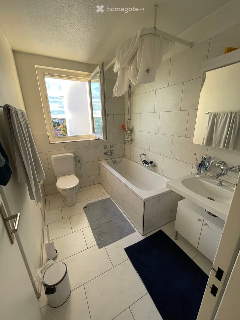 Modern bathroom with white tiles, a bathtub, toilet, sink, and a large window offering a view. Includes towels, a bathmat, and a trash can.