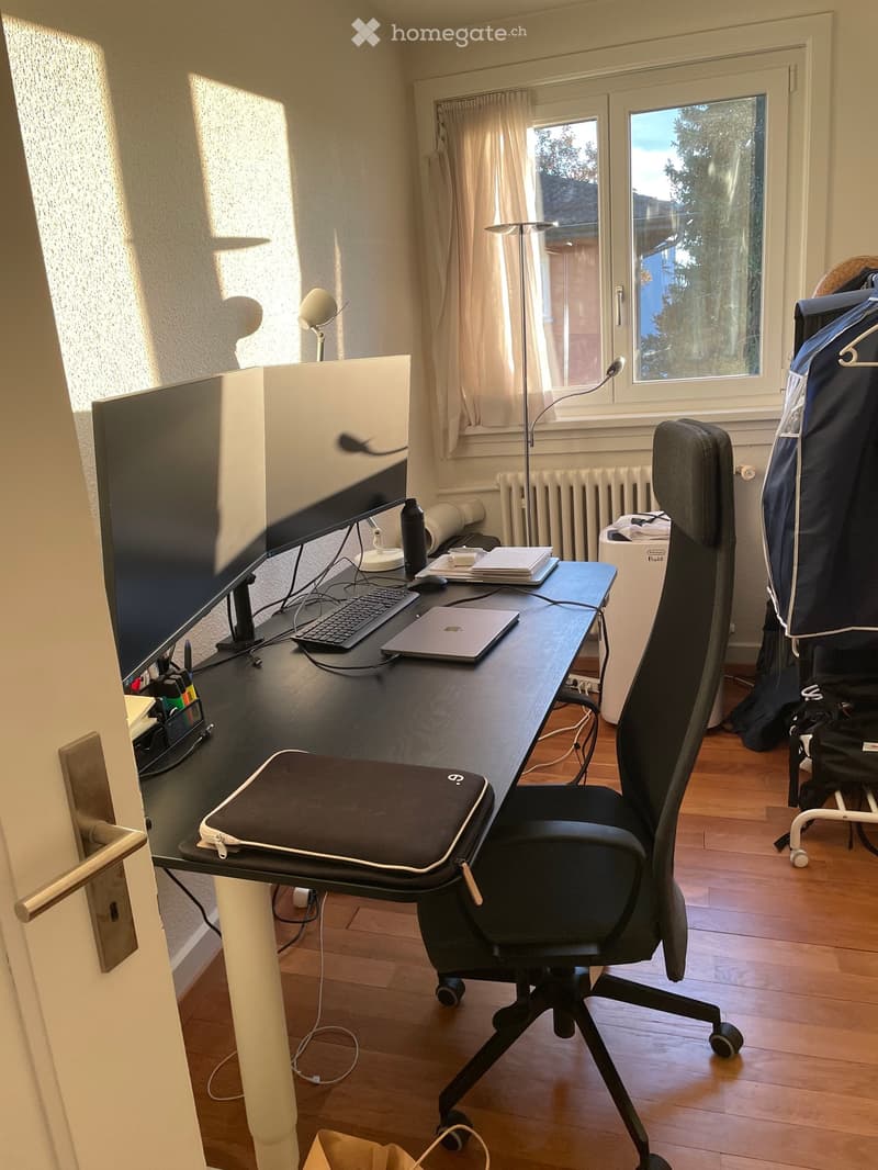 A well-lit room with a wooden floor, a large desk with a laptop, laptop charger, laptop mouse, laptop keyboard, laptop screen, laptop stand, laptop bag, and a laptop sleeve. A black office chair is positioned in front of the desk. There is a window with w