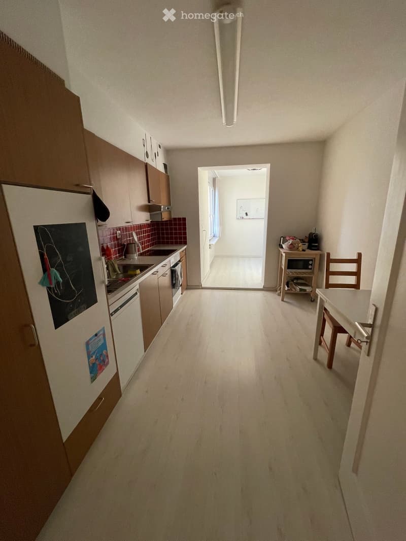 A narrow hallway with light-colored walls and a wooden floor, leading to a kitchen area with cabinets, a sink, and a small dining area with a table and chairs.