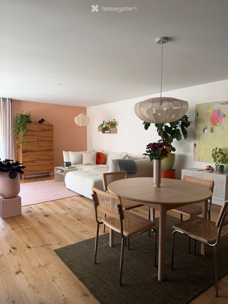Modern living room with a white sofa, round wooden dining table, wicker chairs, pendant light, and decorative plants.