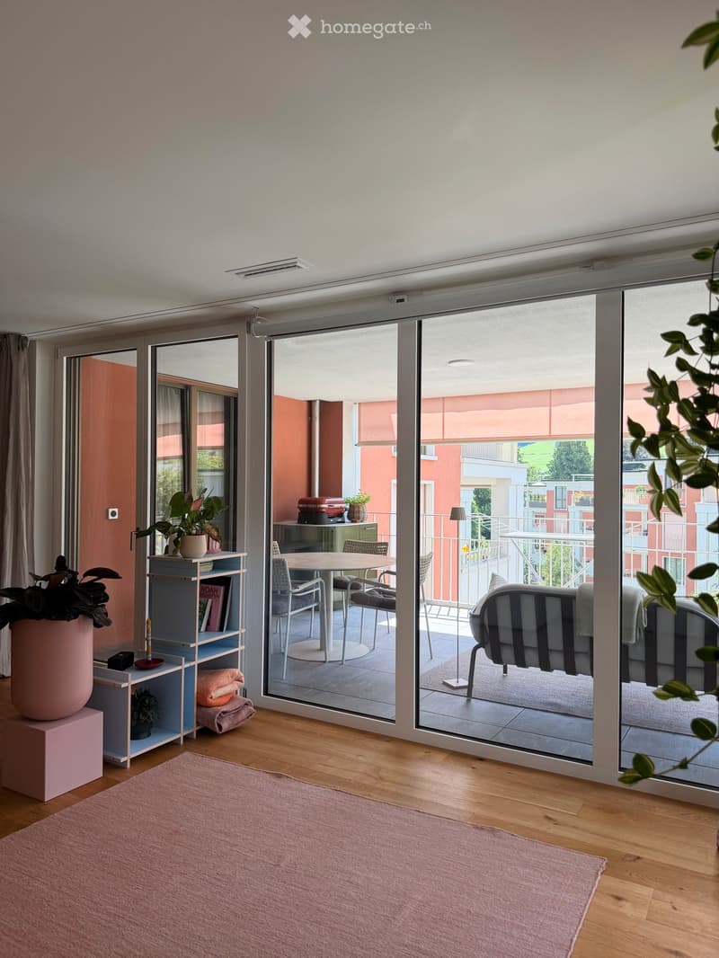 Bright living room with wooden flooring, large sliding glass doors opening to a balcony, sofa, dining table, chairs, shelf with books and plants, and a pink rug.