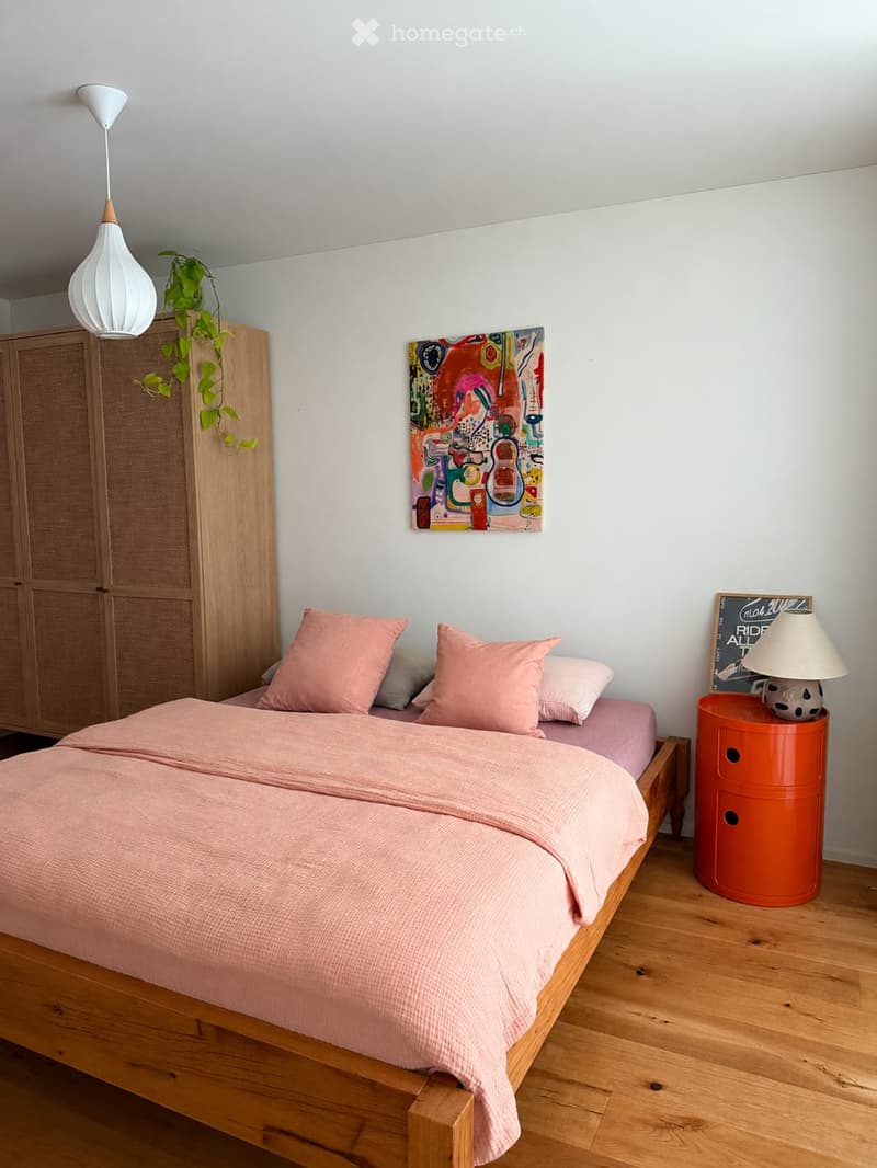 A modern bedroom with a wooden bed, pink bedding, a nightstand with a lamp, a wooden cabinet, a colorful painting, and a hanging plant.
