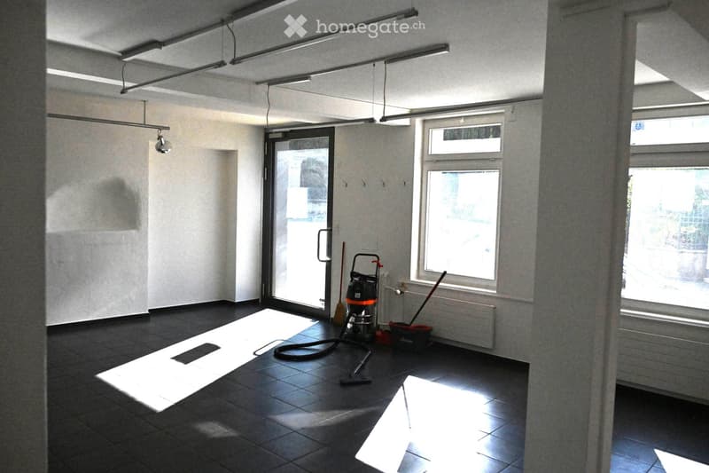 Empty room with dark tiled floor, white walls, large windows, and a glass door leading outside. Cleaning tools are placed near the door.