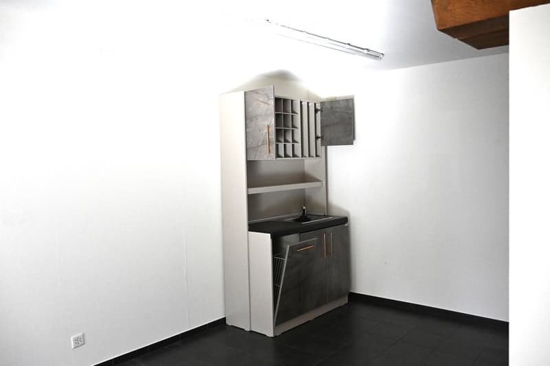Modern kitchen with sleek, grey cabinets, a black countertop, and a built-in sink. The space is clean and minimalistic with white walls and dark tiled flooring.