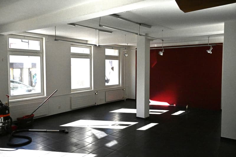 Empty room with dark tiled floor, white walls, large windows, and a red accent wall. No furniture or equipment present.