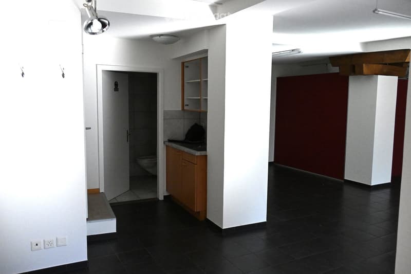 Empty room with black tiled floor, white walls, and a small kitchen area with wooden cabinets and a countertop. A doorway leads to a bathroom with a toilet and a shower stall.