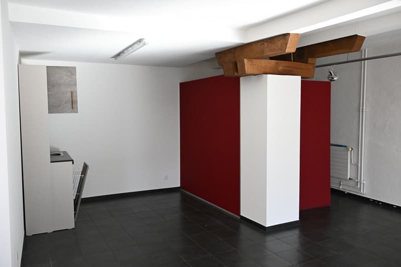 Empty room with white walls, dark tiled floor, and a red accent wall. Exposed wooden beams on the ceiling and a small counter with a sink are visible.