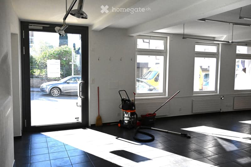 Empty room with tiled floor, large windows, and cleaning equipment near the entrance.