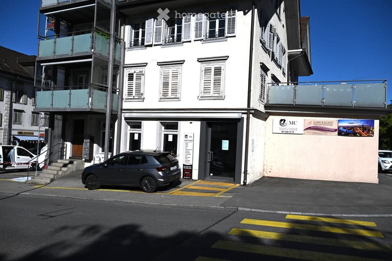 Multi-story building with white facade, multiple windows, and glass balconies. A storefront with signage is visible on the ground floor, and a car is parked in front.