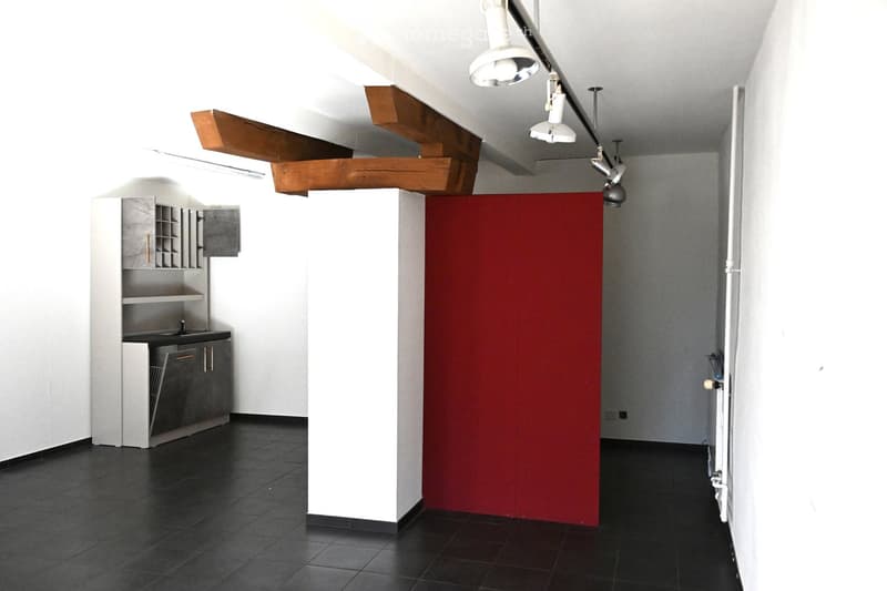 Modern, minimalist room with white walls, dark tiled floor, and a red partition. Features built-in shelving and a small sink area, with exposed wooden beams on the ceiling and track lighting.