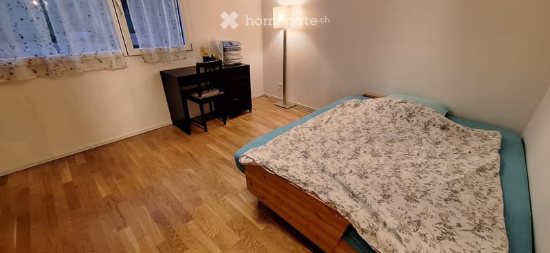 Cozy bedroom with wooden flooring, a single bed with a patterned duvet, a desk with a chair, and a floor lamp near the window.