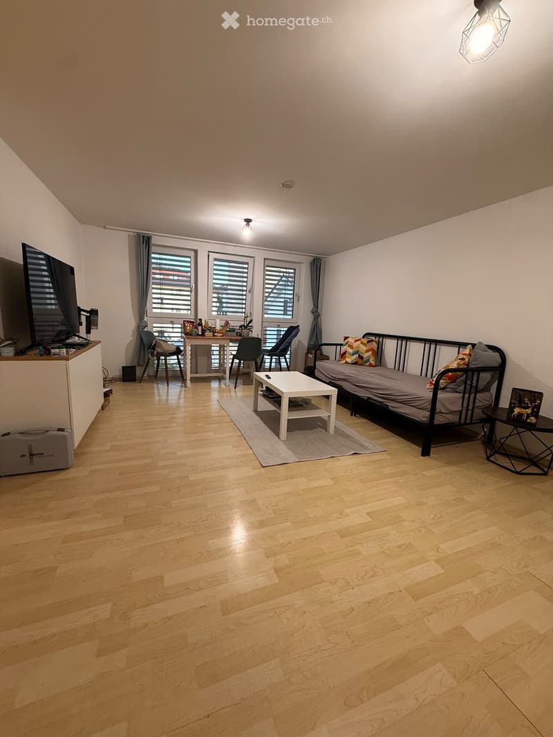 A modern, multi-functional room with a sofa bed, coffee table, small table, chairs, and a TV screen. The room features wooden flooring, large windows with blinds, and a cozy layout.
