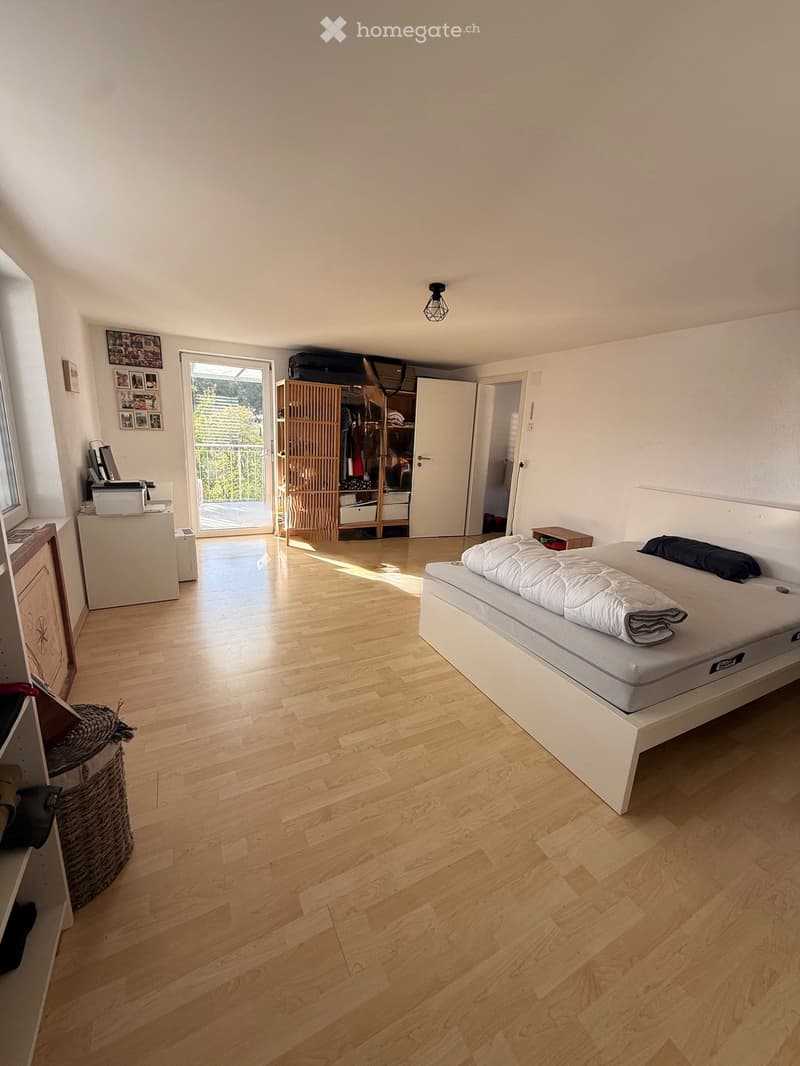 Bright bedroom with wooden flooring, a white bed with a quilt, a wicker basket, a wardrobe, and a printer on a small table. A sliding glass door leads to a balcony.