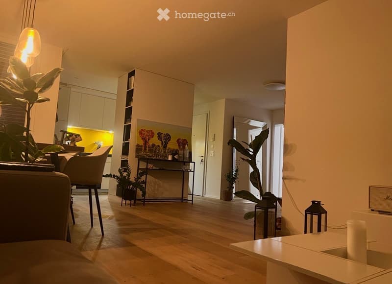 Modern living room with wooden flooring, white walls, and large windows. Features a sofa, chair, small table, shelf with decorative items, potted plants, and a picture frame on the wall.
