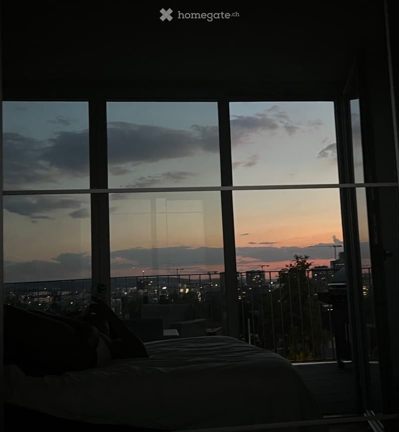 Bedroom with large windows offering a cityscape view at sunset, featuring a bed and a chair.