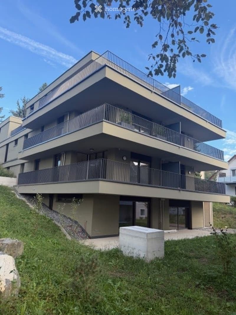 Modern multi-story residential building with balconies, large windows, and a raised ground floor on a grassy slope.