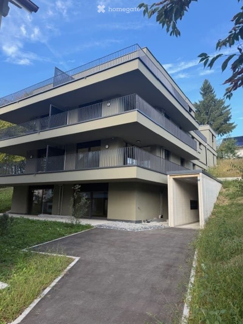 Modern multi-story residential building with clean lines, large windows, and balconies. The structure is set on a hillside with a paved driveway leading to the entrance.