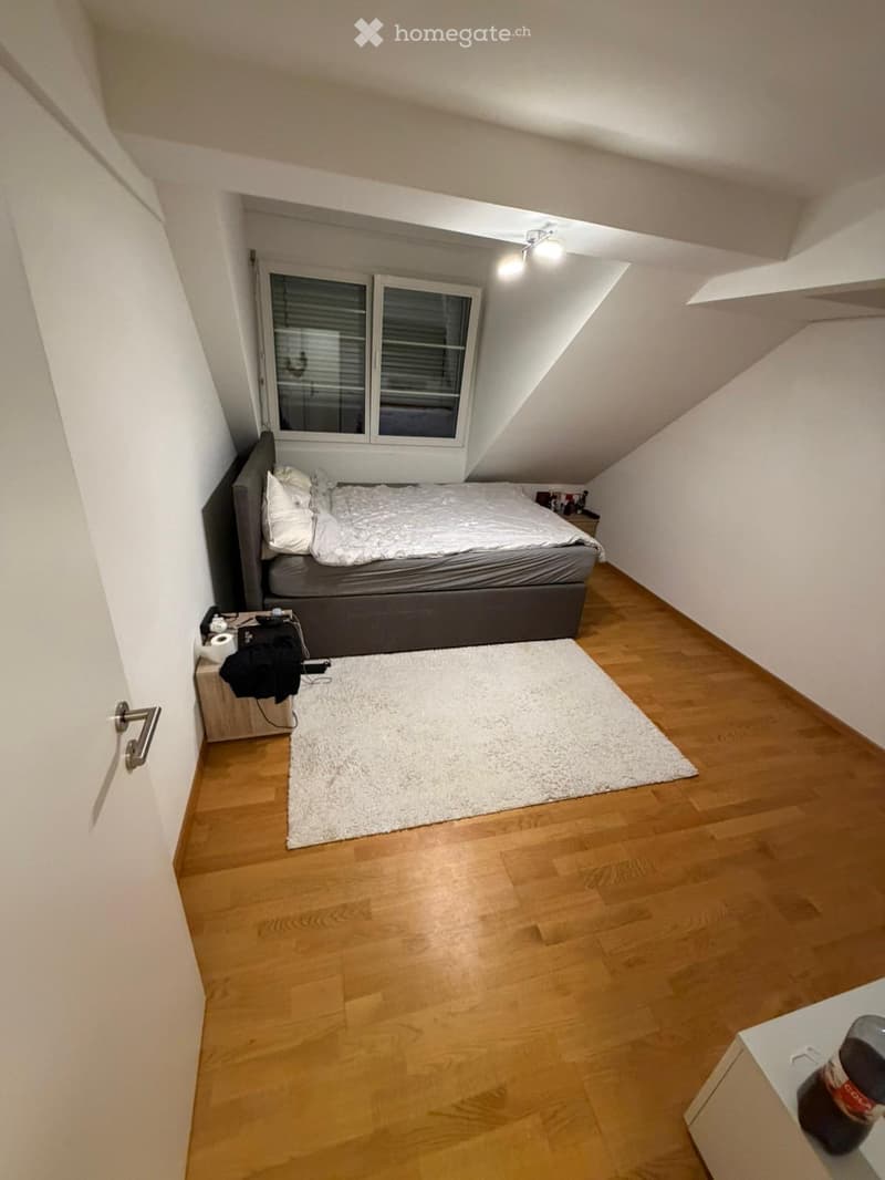 Cozy attic bedroom with wooden flooring, a large bed with white bedding, a nightstand, and a white rug.