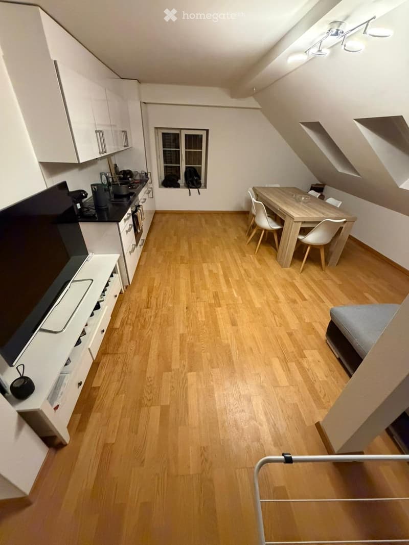 A modern, compact kitchen and living area with light wooden flooring, white cabinets, a stove, refrigerator, dining table with chairs, and a small sofa.
