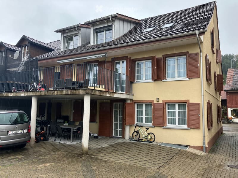 Two-story house with beige exterior, multiple windows with shutters, garage, and balcony with outdoor furniture.