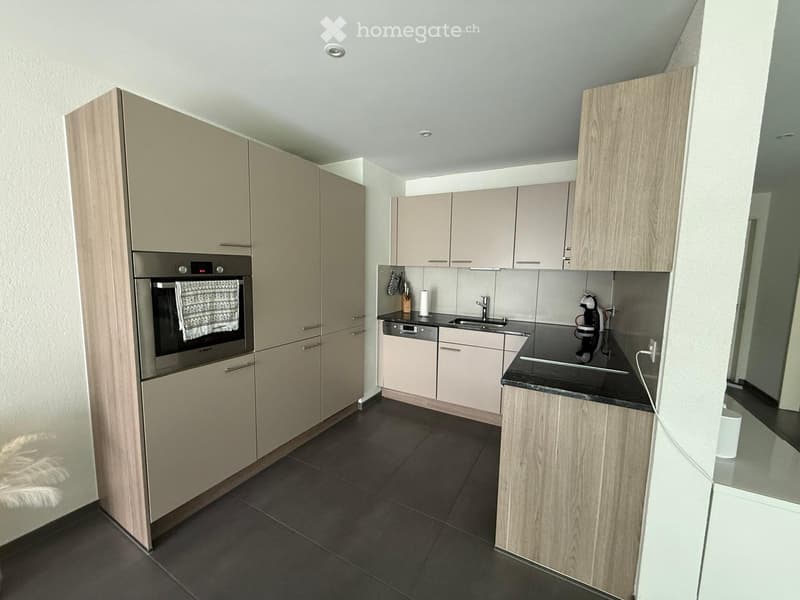 Modern kitchen with light wood cabinets, stainless steel appliances, black countertops, and dark tiled flooring.