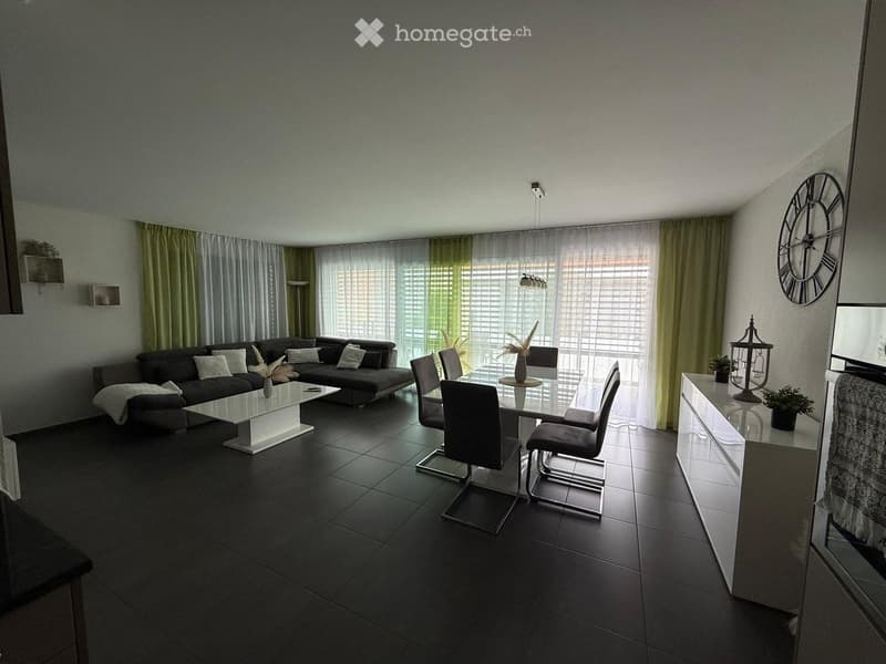 Modern living and dining area with dark grey sofa, white coffee table, glass dining table with chairs, large windows with curtains, and decorative items including a clock and sideboard.