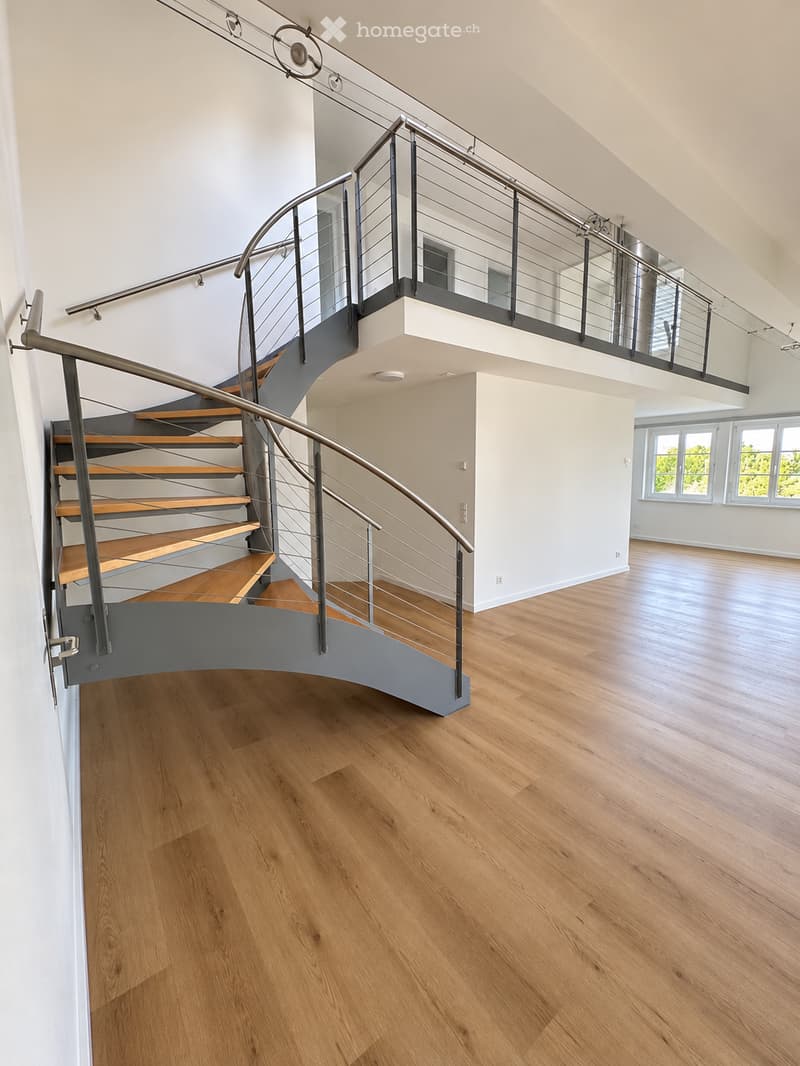 Modern interior hallway with wooden flooring, a curved staircase with metal railings, and large windows allowing natural light to enter.