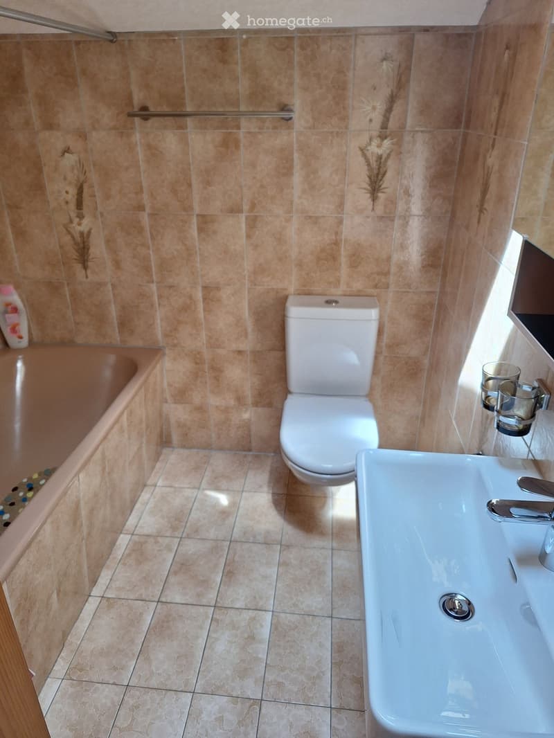 Bathroom with beige tiled walls and floor, featuring a toilet, sink, bathtub, and a small shelf with cups.
