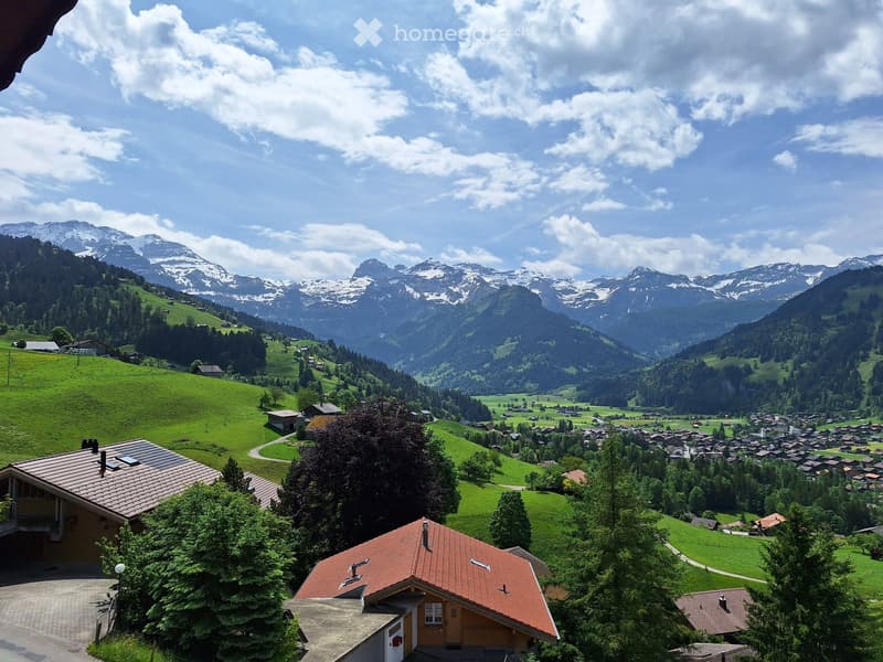 Panoramic view of a village nestled in a lush green valley surrounded by snow-capped mountains. The foreground shows a house with a red-tiled roof and a driveway, while the background features rolling hills, scattered houses, and a winding road.