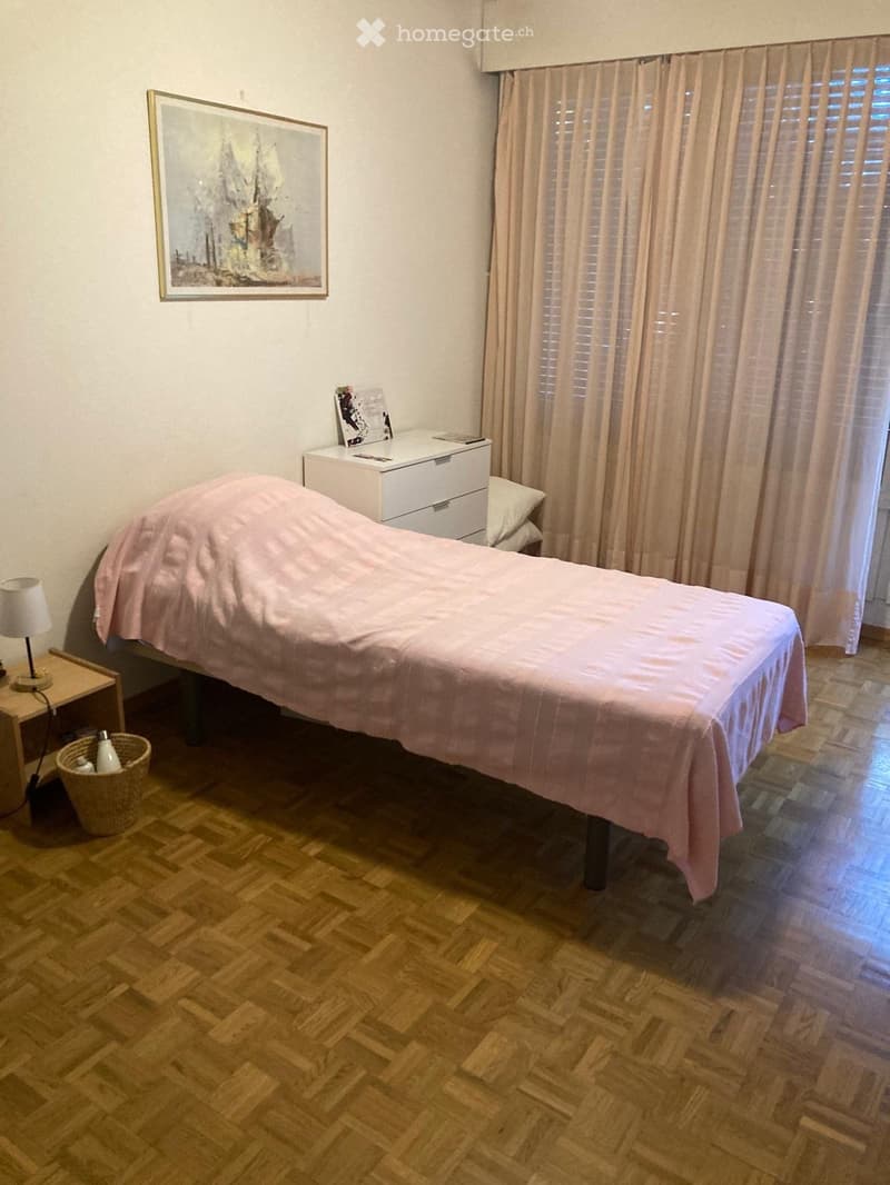 Cozy bedroom with wooden herringbone flooring, a single bed with pink bedding, a nightstand with a lamp, a drawer unit, and a wicker basket on the floor.