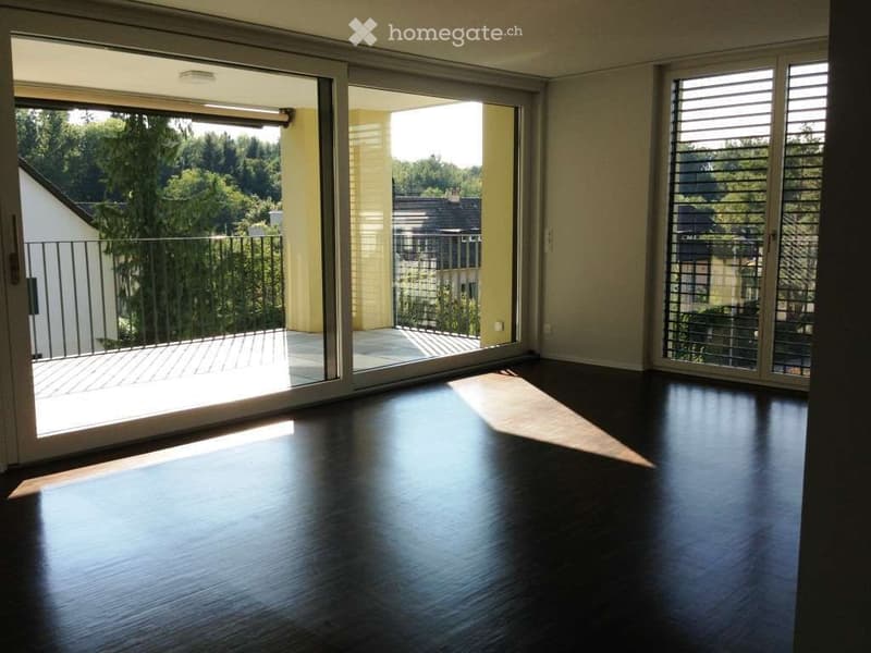 Empty room with dark wooden flooring, large sliding glass doors leading to a balcony with a metal railing, and a view of greenery and neighboring buildings.