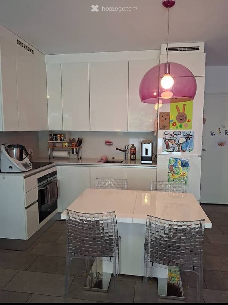 Modern kitchen with white cabinets, a central island table with four transparent chairs, a refrigerator with children's drawings, and various appliances including a blender and oven.
