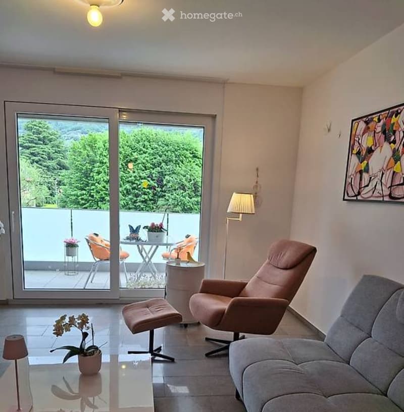 Modern living room with large sliding glass doors leading to a balcony, featuring a grey sofa, leather chair, small round table, and decorative flower pots.