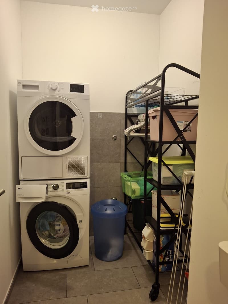Laundry area with stacked washing machine and dryer, storage rack with bins, and a blue trash bin.