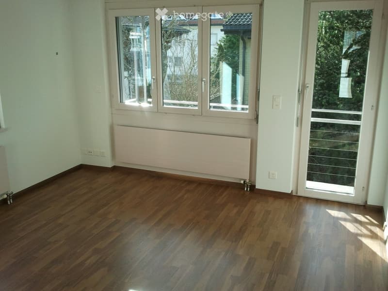 Empty room with wooden flooring, large windows, radiator, and sliding glass door leading outside.