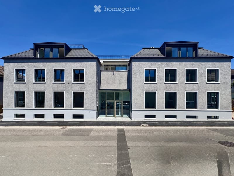 Modern two-story building with light gray facade, multiple windows, and a central glass entrance. The roof features dormer windows and a small balcony area.