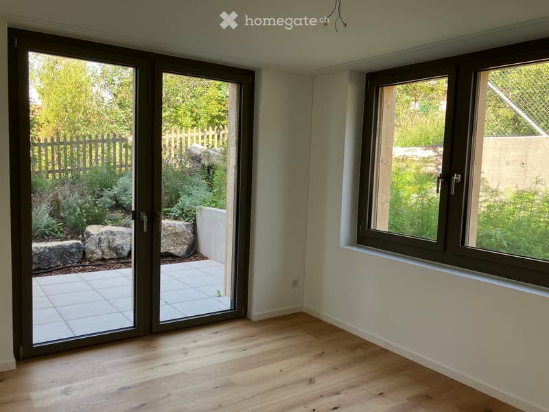 Empty room with wooden flooring, large sliding glass doors leading to a garden, and a window with a view of greenery.