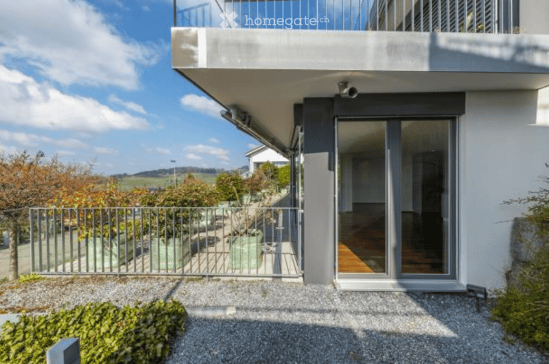 Modern two-story house with a raised ground floor, large balcony, and contemporary design. The entrance features large glass doors, and the surrounding area includes a gravel driveway, a metal gate, and landscaped greenery.