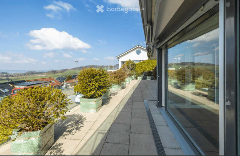 Balcony with tiled floor, potted plants, large glass sliding doors, scenic countryside view.