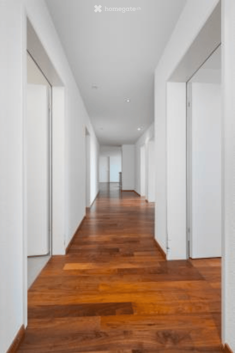 Long, narrow hallway with polished wooden flooring and white walls, featuring recessed lighting and closed doors on both sides.