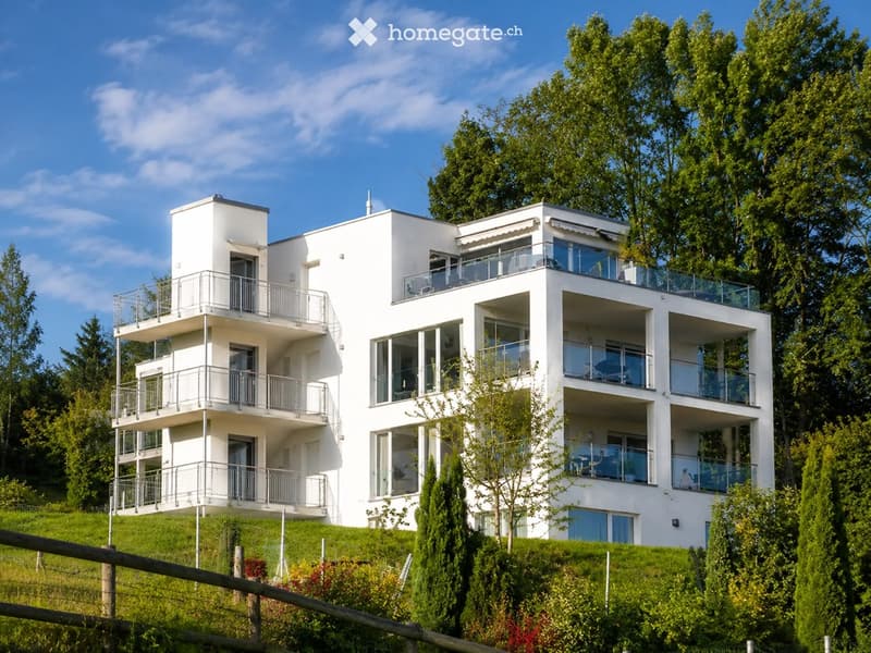 Modern white multi-story building with balconies and large windows, surrounded by greenery and trees.