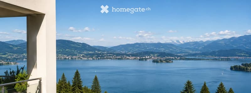 Balcony with panoramic view of a lake and surrounding mountains, clear blue sky, distant town, and trees on the foreground.