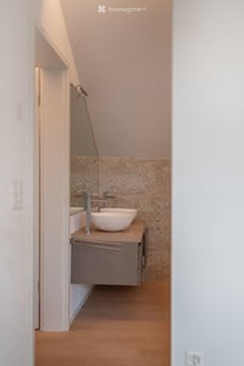 Modern bathroom with a white sink, mirror, and wooden cabinet, featuring a sloped ceiling and light-colored tiles.