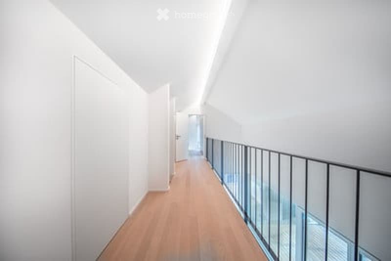 Bright, modern hallway with wooden flooring, white walls, and a glass railing leading to an open area.