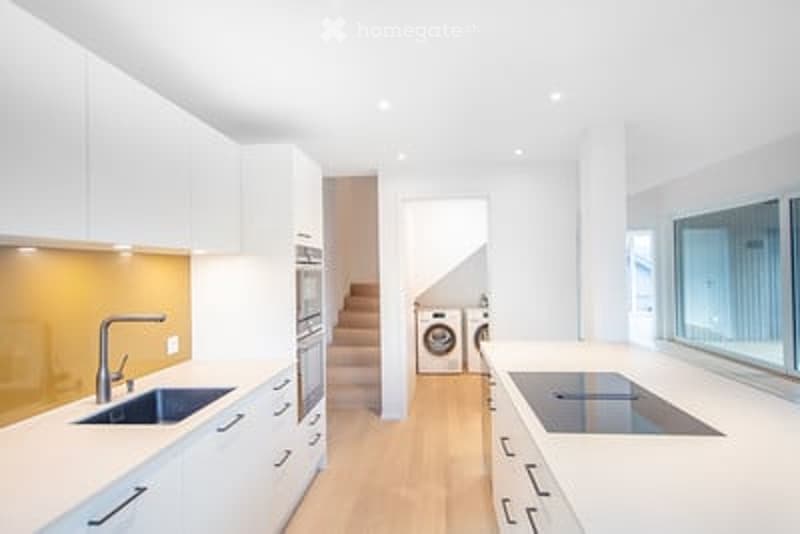 Modern, minimalist kitchen with white cabinets, stainless steel appliances, and a central island with a built-in stove. Features a sink with a modern faucet, and a washing machine is visible in the adjacent space.