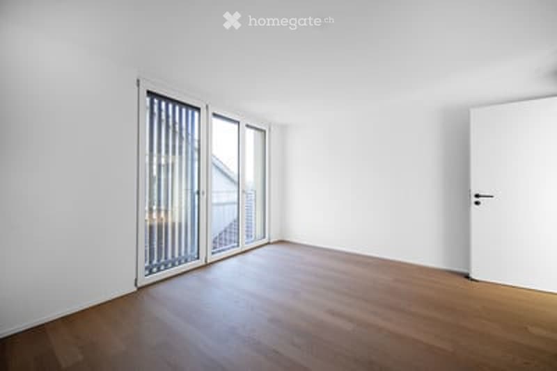 Completely empty room with white walls, wooden floor, large sliding glass doors, and a white door.