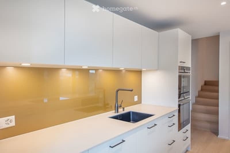 Modern kitchen with white cabinets, a stainless steel sink, and a built-in oven. The backsplash features a warm, golden glass design.