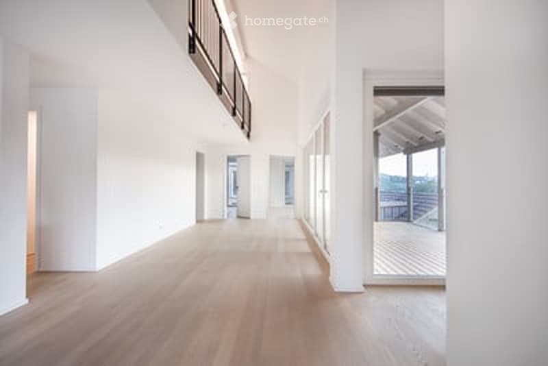 Bright, open-plan interior with light wooden flooring and white walls. Features large windows and a balcony area visible at the end of the corridor.