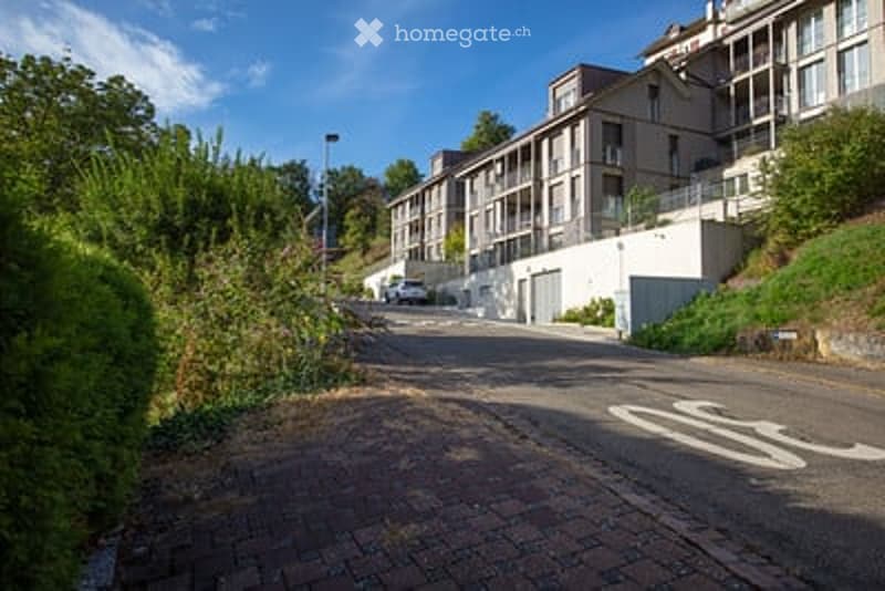 A row of modern apartment buildings on a hillside, with balconies and parking areas visible.